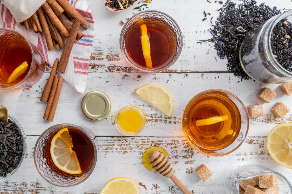 Vista superior de copos de chá com limão, paus de canela, mel, açúcar mascavo e folhas de chá soltas em uma mesa branca rústica, destacando uma variedade de ingredientes para chás.