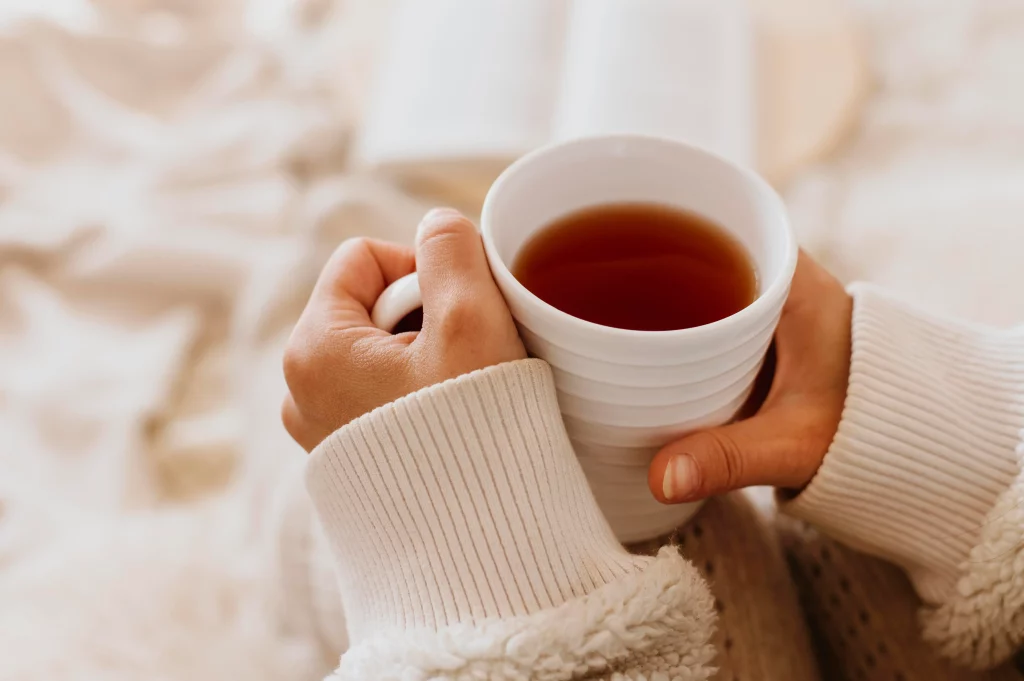 Pessoa segurando uma xícara de chá quente, vestindo um suéter de lã, com um livro desfocado ao fundo, criando um ambiente aconchegante de inverno.