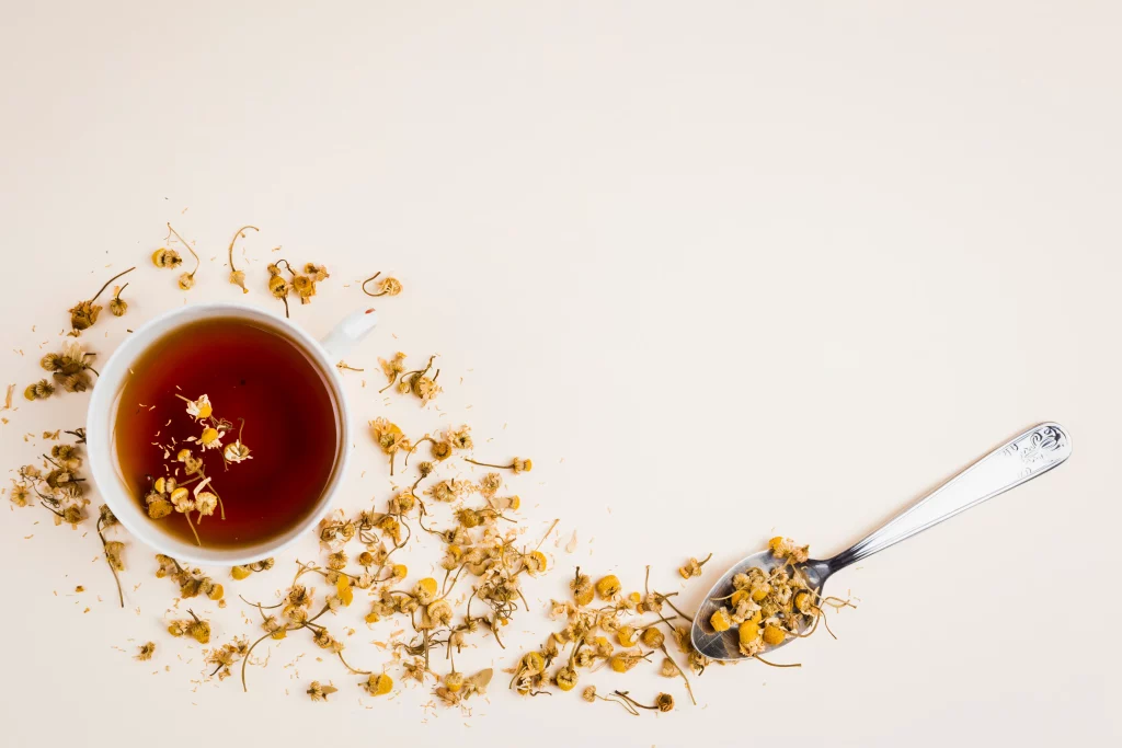 Xícara de chá de camomila cercada por flores de camomila secas e uma colher cheia de flores de camomila, destacando os ingredientes naturais.