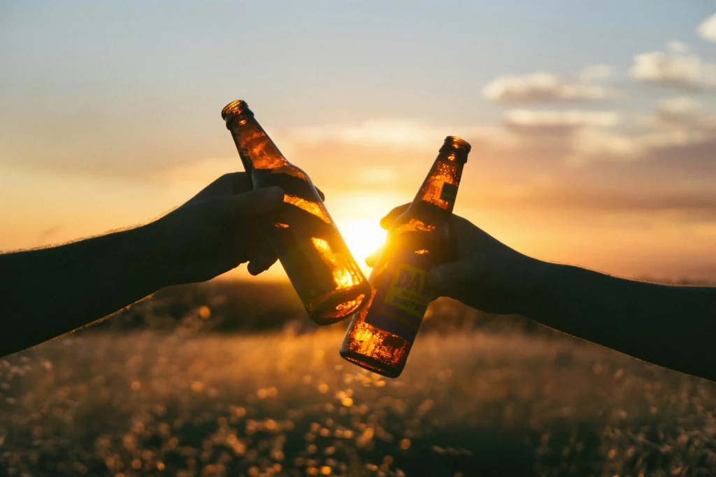 Duas pessoas brindando com garrafas de cerveja ao pôr do sol, com luz do sol brilhando através das garrafas.