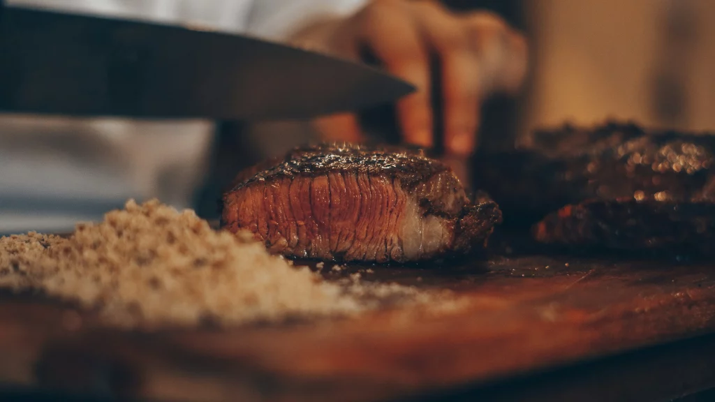 Corte de carne grelhada sendo fatiado com faca, mostrando o ponto ideal de cozimento com interior rosado e exterior selado. Farofa ao lado para acompanhamento, destacando um preparo típico de churrasco.