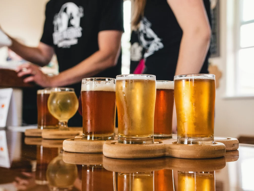 Várias cervejas artesanais em copos sobre uma mesa de madeira, com pessoas ao fundo.