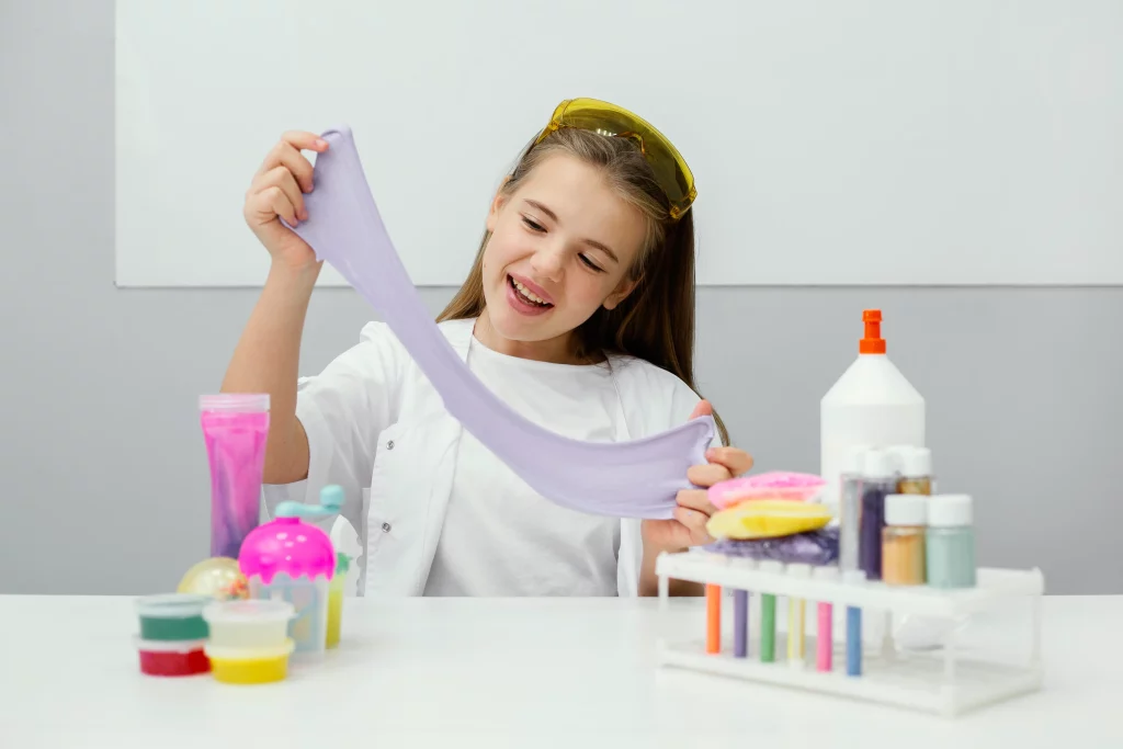 Menina sorridente brinca com um slime roxo, esticando-o com as mãos. Na mesa à sua frente, há materiais de artesanato como cola, corantes e glitter.