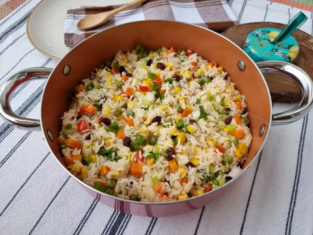 Panela com arroz à grega, colorido com legumes como cenoura, ervilha, pimentão, milho e uva-passa.