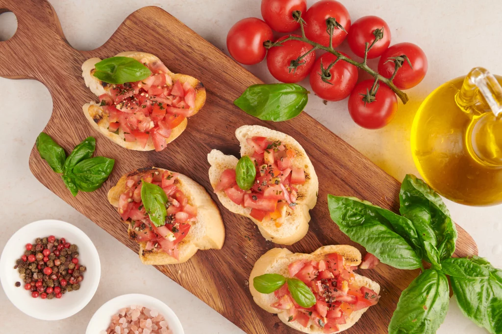 Tábua de madeira com bruschettas de tomate e manjericão, decorada com ingredientes como tomate, folhas de manjericão, azeite e pimentas.