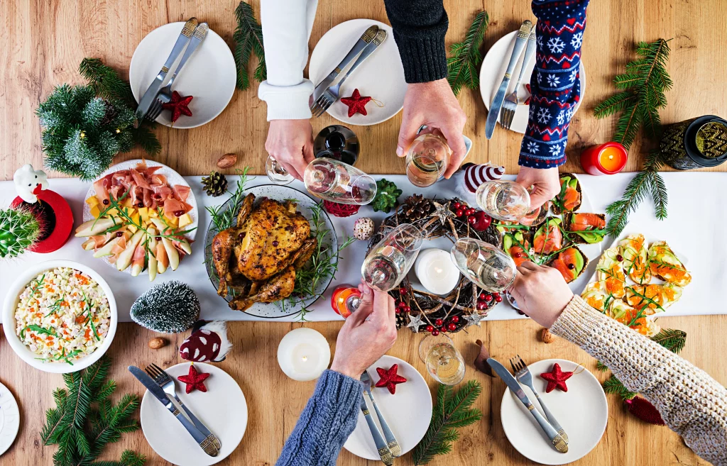 Mesa decorada para ceia de Natal com pratos variados, incluindo frango assado, tábua de frios, salada e aperitivos; mãos brindando com taças de espumante, ao redor de uma decoração festiva com velas e enfeites natalinos.