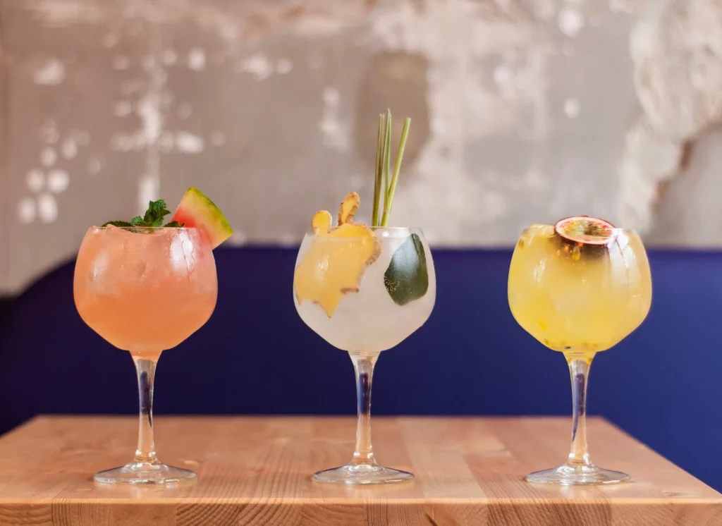 Três drinks coloridos servidos em taças de gin, decorados com frutas e ervas. 