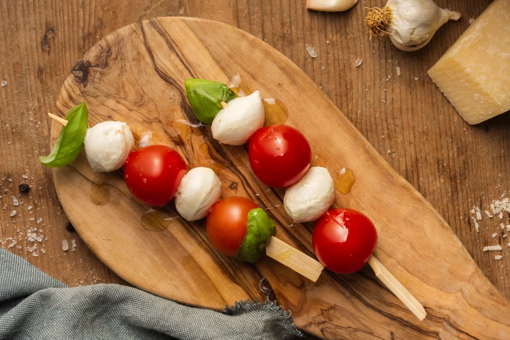 Espetinho caprese com tomate cereja, mussarela de búfala e folhas de manjericão, servido em uma tábua de madeira.