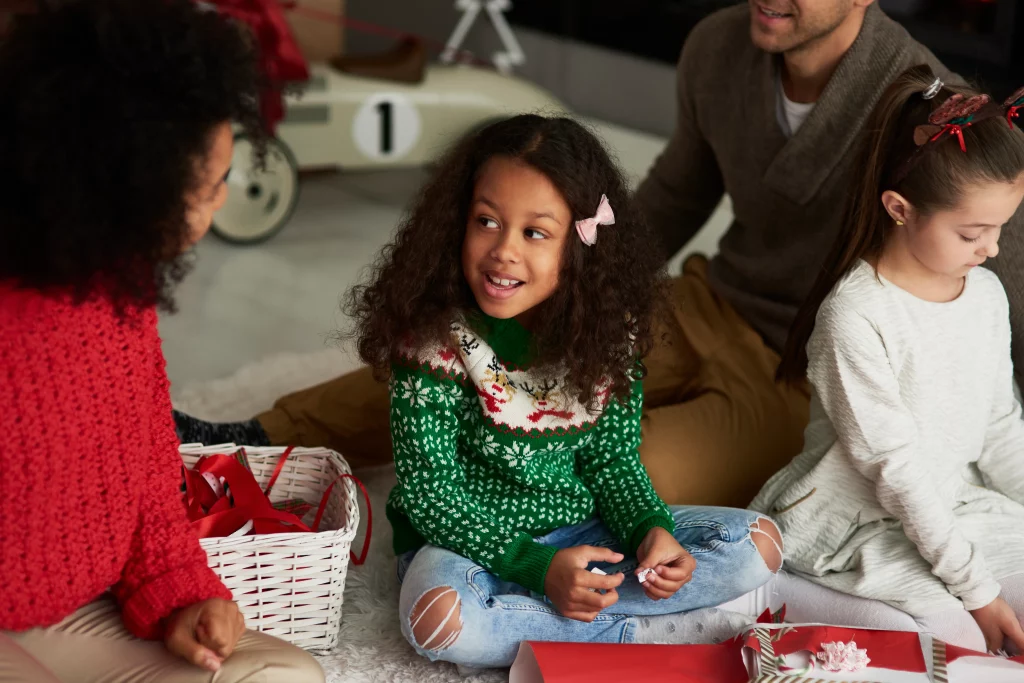 Família reunida empacotando presentes com uma cesta de Natal ao fundo, incluindo crianças sorrindo e participando da decoração.