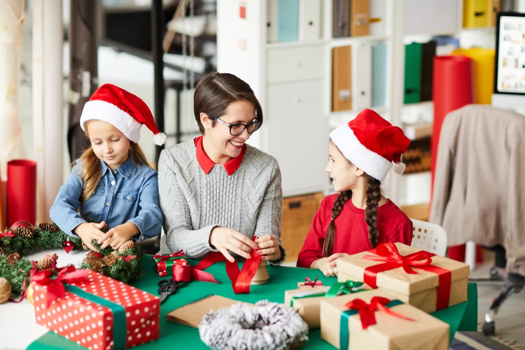 Mãe e duas filhas com gorros de Papai Noel preparando presentes de Natal com fitas e papéis decorativos em uma mesa.