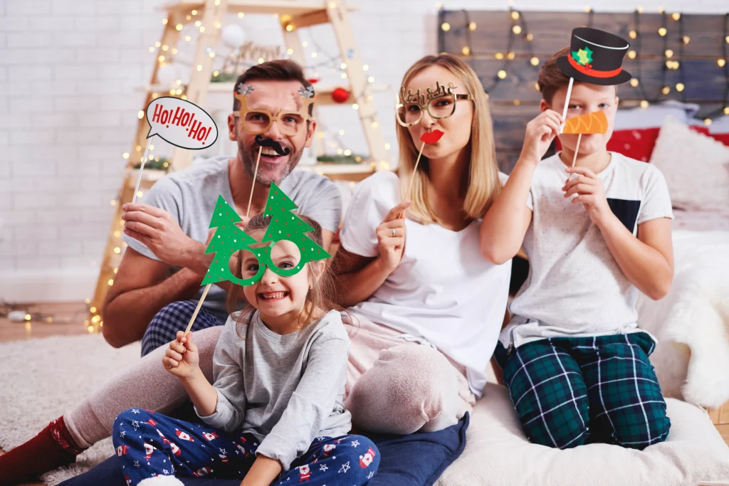 Família reunida posando com acessórios natalinos, como óculos, chapéus e plaquinhas decorativas, em uma sala com decoração de Natal.