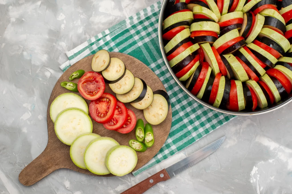 Preparação de Ratatouille com fatias de abobrinha, berinjela, tomate e pimenta, dispostas em uma travessa e em uma tábua de madeira.