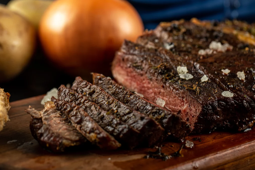 Close-up de um suculento bife grelhado fatiado, temperado com ervas e sal grosso, sobre uma tábua de madeira, com cebolas e batatas desfocadas ao fundo.