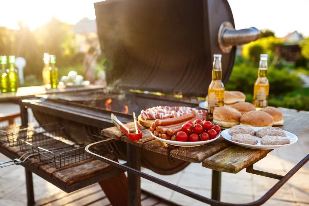 Churrasco com carne, salsicha e hambúrguer pronto para assar, acompanhado de bebidas e pães, em um dia ensolarado.