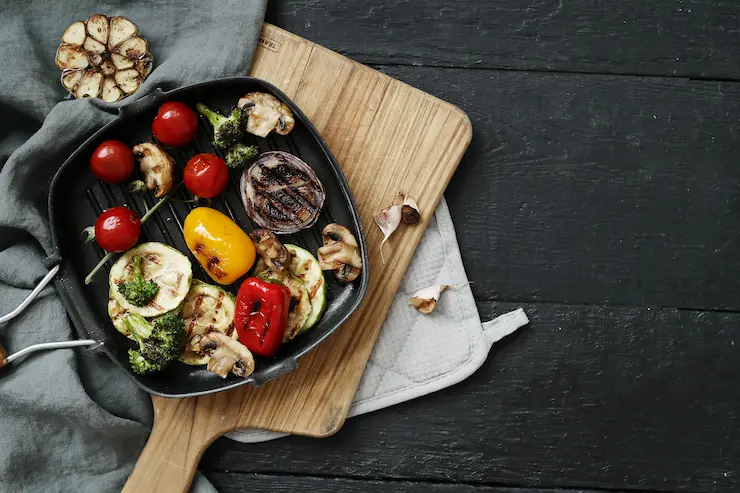 Prato de legumes grelhados variados como pimentão, tomate, brócolis e cogumelos, servidos como acompanhamentos para churrasco.